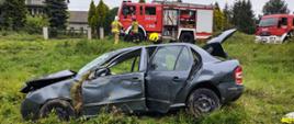 Samochód osobowy na łące z podniesioną klapą bagażnika. Na drodze samochody pożarnicze oraz strażacy.