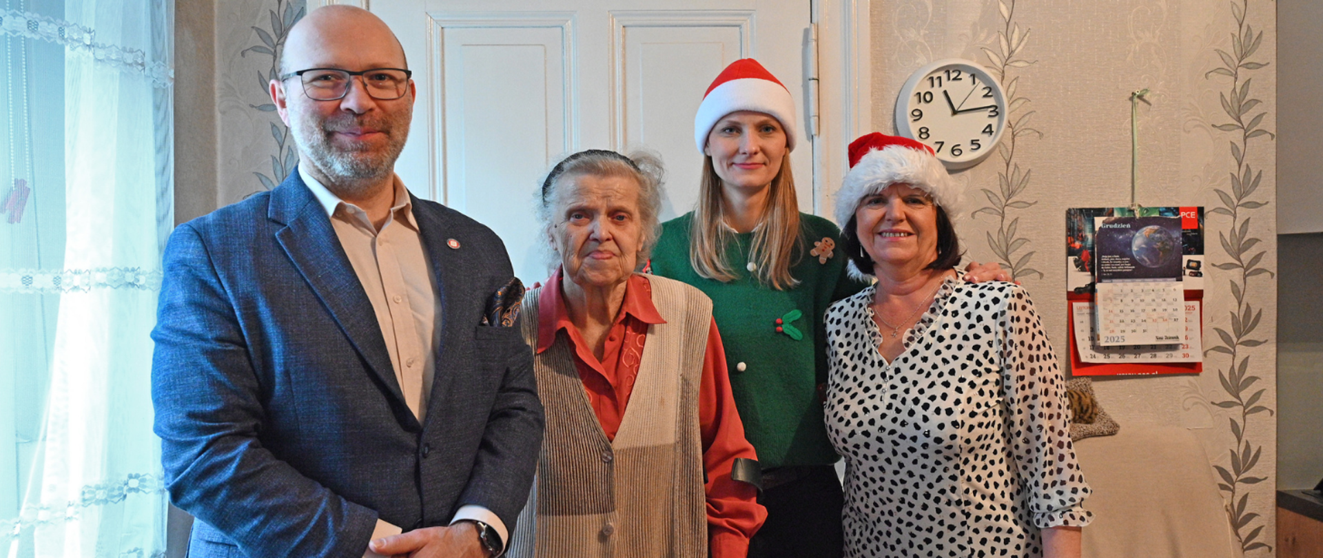 Cztery osoby stojące w mieszkaniu w świątecznej atmosferze. W centrum starsza kobieta, obok niej trzy osoby – dwie kobiety w czapkach świątecznych oraz mężczyzna w marynarce. W tle widoczny zegar ścienny i kalendarz. Zdjęcie przedstawia spotkanie związane z przekazaniem świątecznej pomocy.
