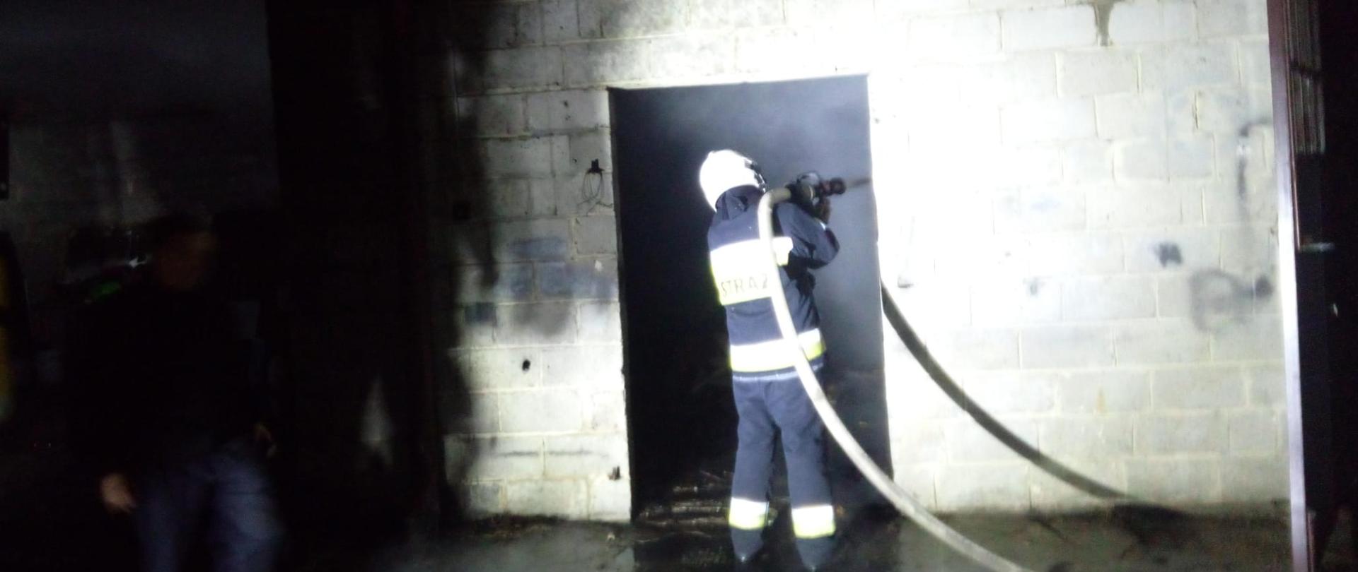 Zdjęcie przedstawia strażaka, który podaje strumień wody do wnętrza budynku gospodarczego gdzie wybuchł pożar.