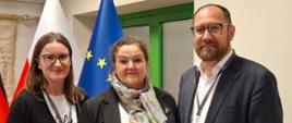 Trzy osoby – dwie kobiety i mężczyzna – stoją obok siebie i pozują do zdjęcia. Kobieta po lewej stronie i mężczyzna po prawej mają okulary. Mężczyzna ma brodę i jest ubrany w ciemny garnitur. Kobieta w środku ma na sobie czarną marynarkę i jasny, wzorzysty szalik. W tle widoczne są flagi Polski i Unii Europejskiej.