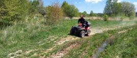 Widok na teren poligonu porośniętego krzewami i wysoką trawą. Pośrodku pojazd marki quad prowadzony przez strażaka.