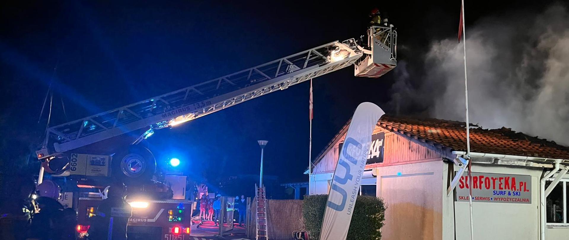 Po prawej palący się parterowy budynek nad który unosi się dym. Po lewej stoi drabina mechaniczna wysuniętymi przęsłami i koszem nad budynkiem