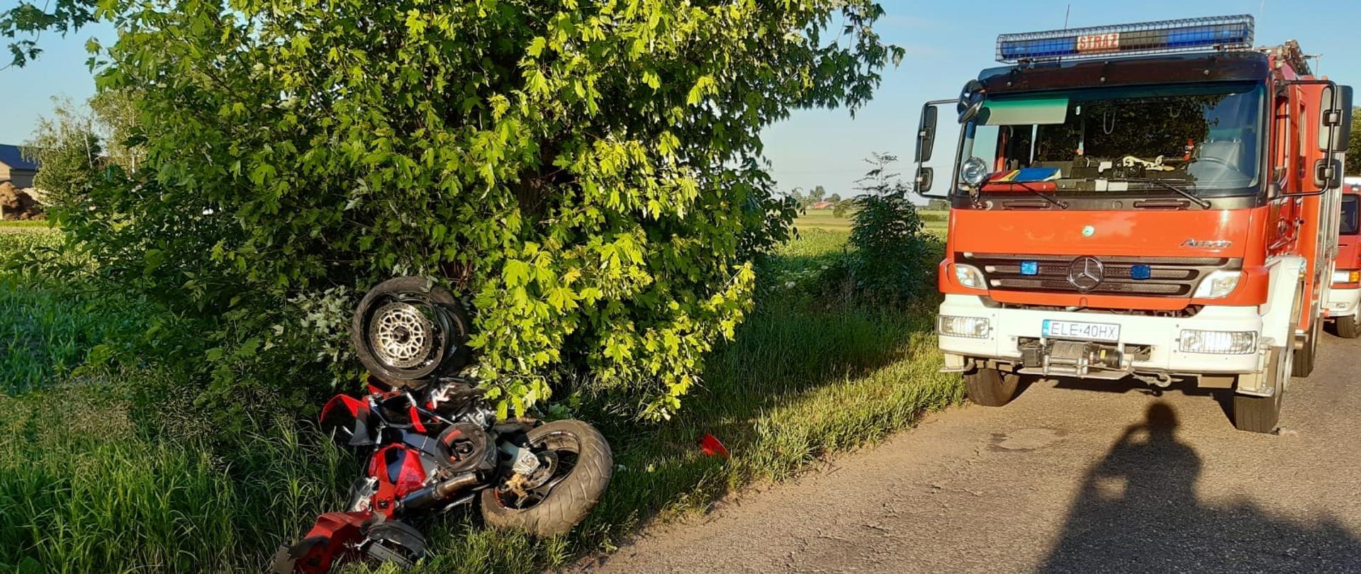 Na zdjęciu widać przewrócony motocykl, który jest w rowie obok drzewa. Na asfaltowej drodze stoi czerwony samochód strażacki ustawiony przodem do motocykla. Za nim widać częściowo drugi samochód strażacki. Jest widno.
