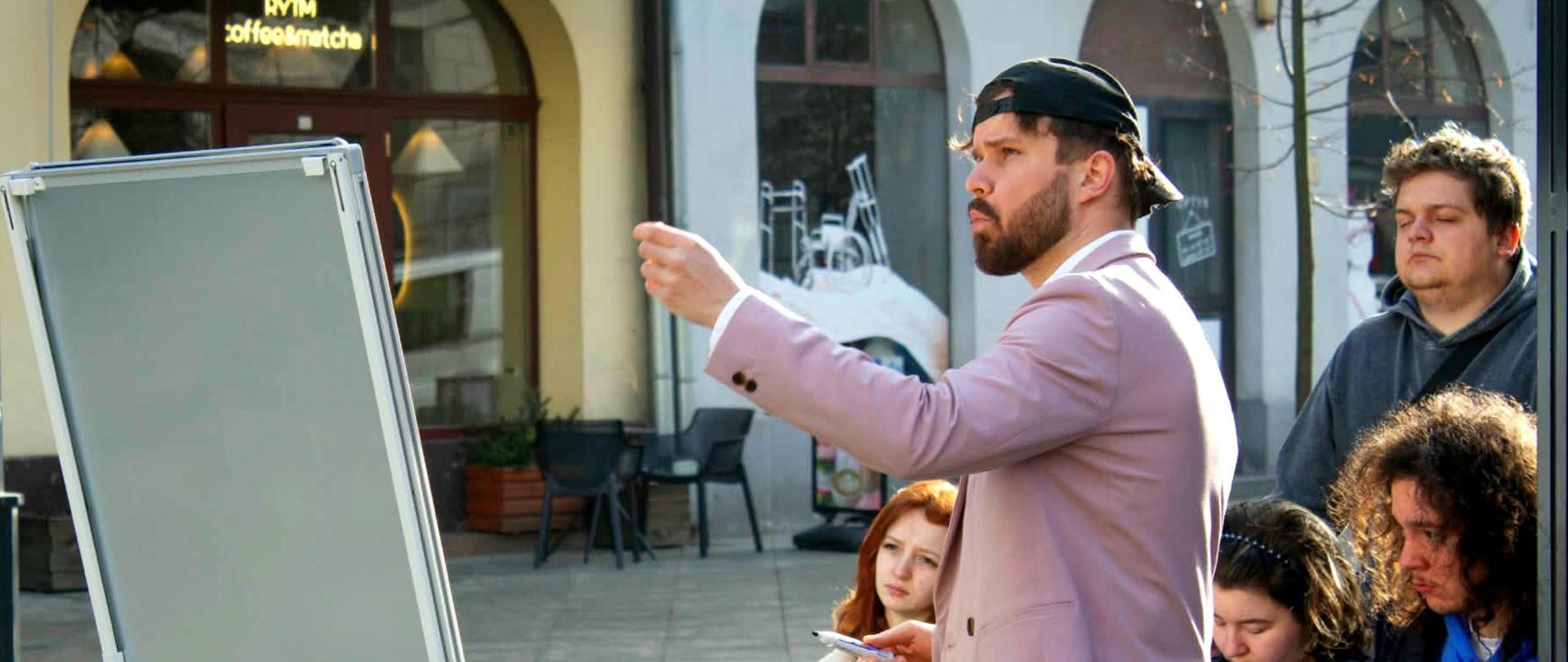 Zajęcia plenerowe z historii sztuki na kaliskim rynku – prowadzący omawia detale architektoniczne przy mobilnej tablicy, a słuchacze notują obserwacje.