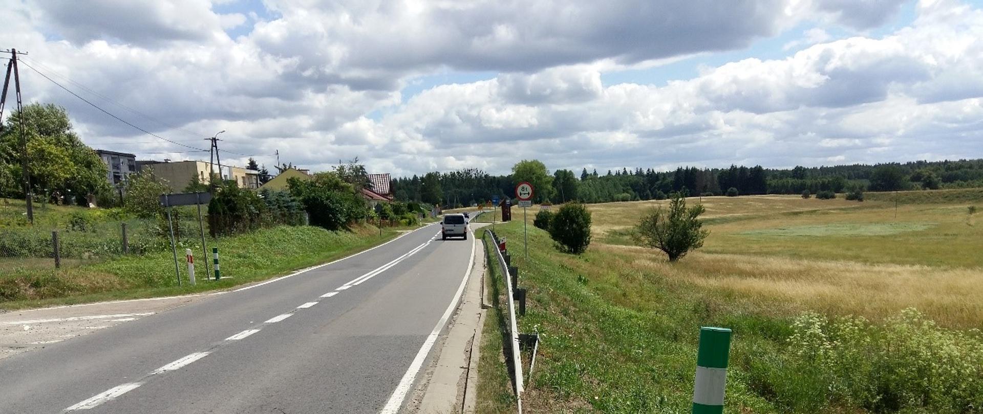 Droga, po której poruszają się samochody przebiega w pobliżu wiejskiej zabudowy jednorodzinnej. Budynki znajdują się z lewej strony drogi, po prawej są łąki, na horyzoncie widoczny jest las.