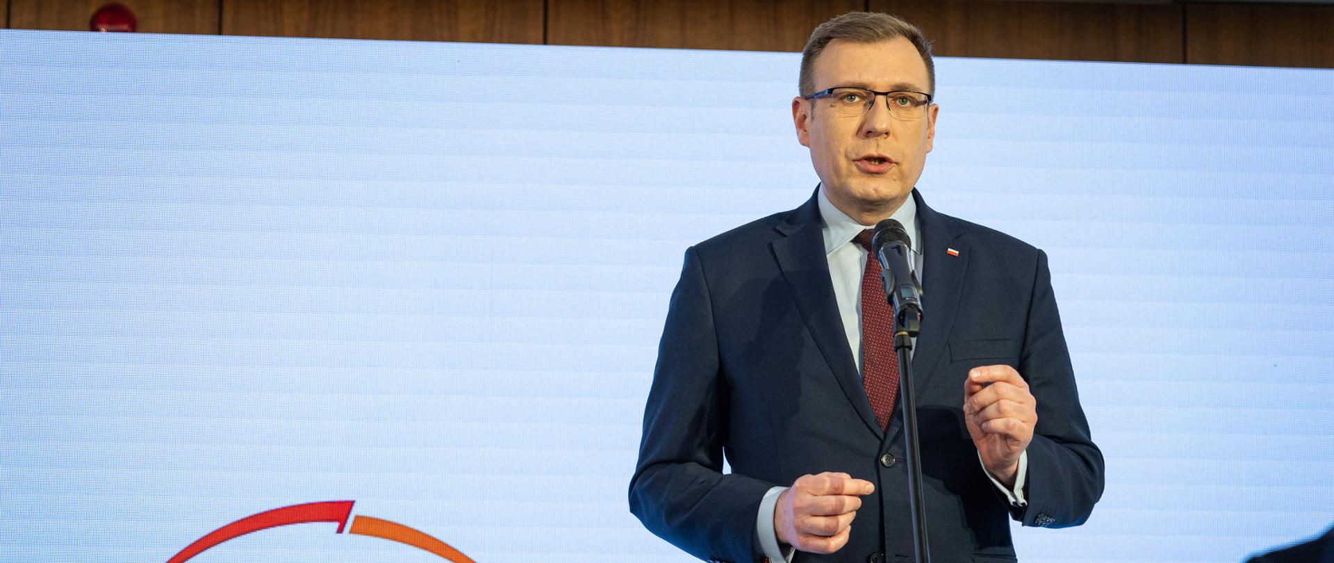 Wiceminister Maciej Małecki stoi za mikrofonem. W tle ekran z logotypem PGE. 