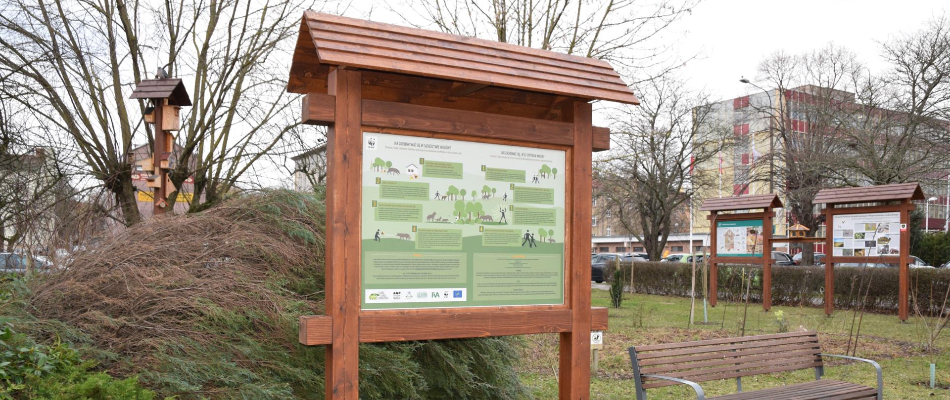 Drewniana tablica stojąca na trawie. Po lewej stronie w oddali słup z budkami dla ptaków, po prawej stronie u dołu stoi ławka, w oddali dwie inne tablice.