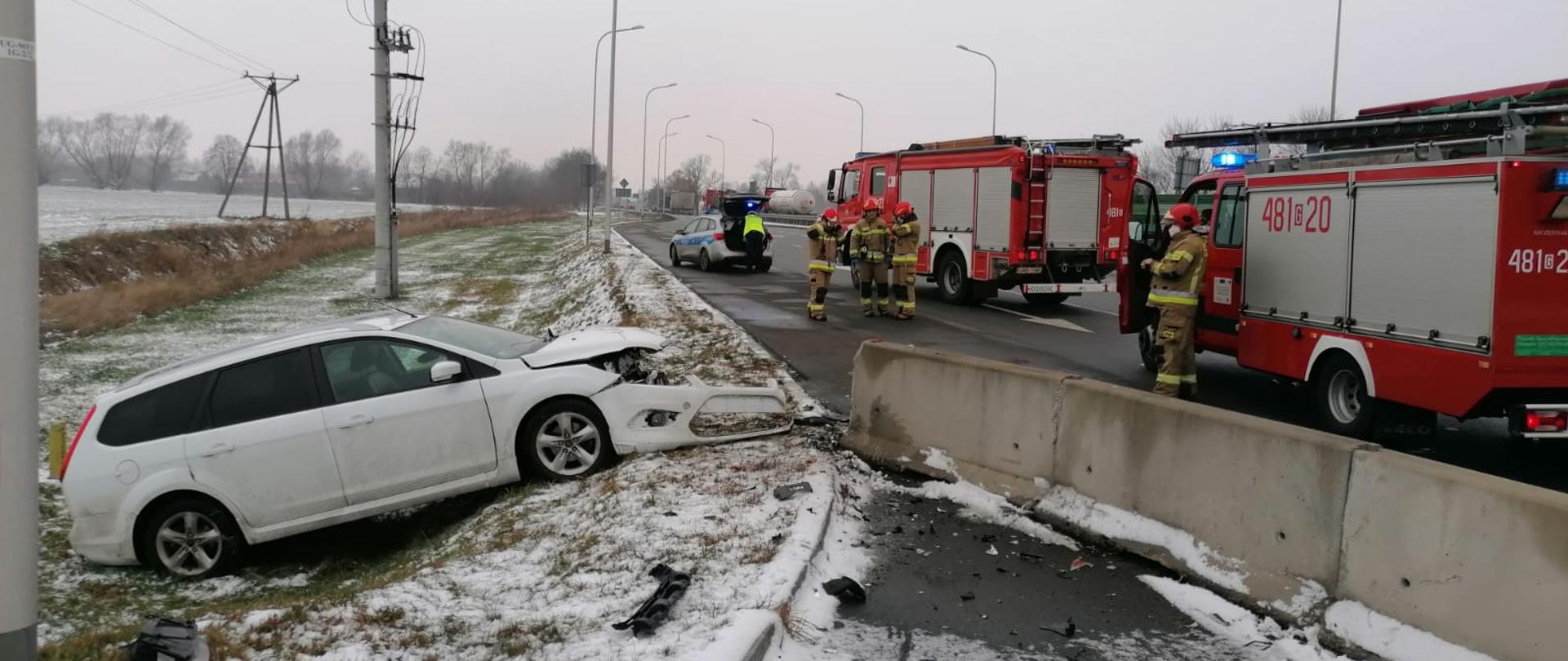 Fotografia przedstawia dwa czerwone samochody strażackie, jeden srebrny patrol policji oraz strażaków i policjantów przy rozbitym samochodzie koloru srebrnego marki ford.