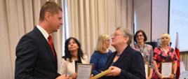 Wiceminister Piontkowski wręcza kobiecie w czarnej marynarce oprawiony w ramki dyplom, za nimi stoi grupa ludzi.