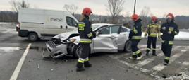 Samochód koloru jasnego biorący udział w zdarzeniu stoi na skrzyżowaniu. Obok niego stoi czterech strażaków. Na dalszym tle samochody uczestniczące w ruchu drogowym..