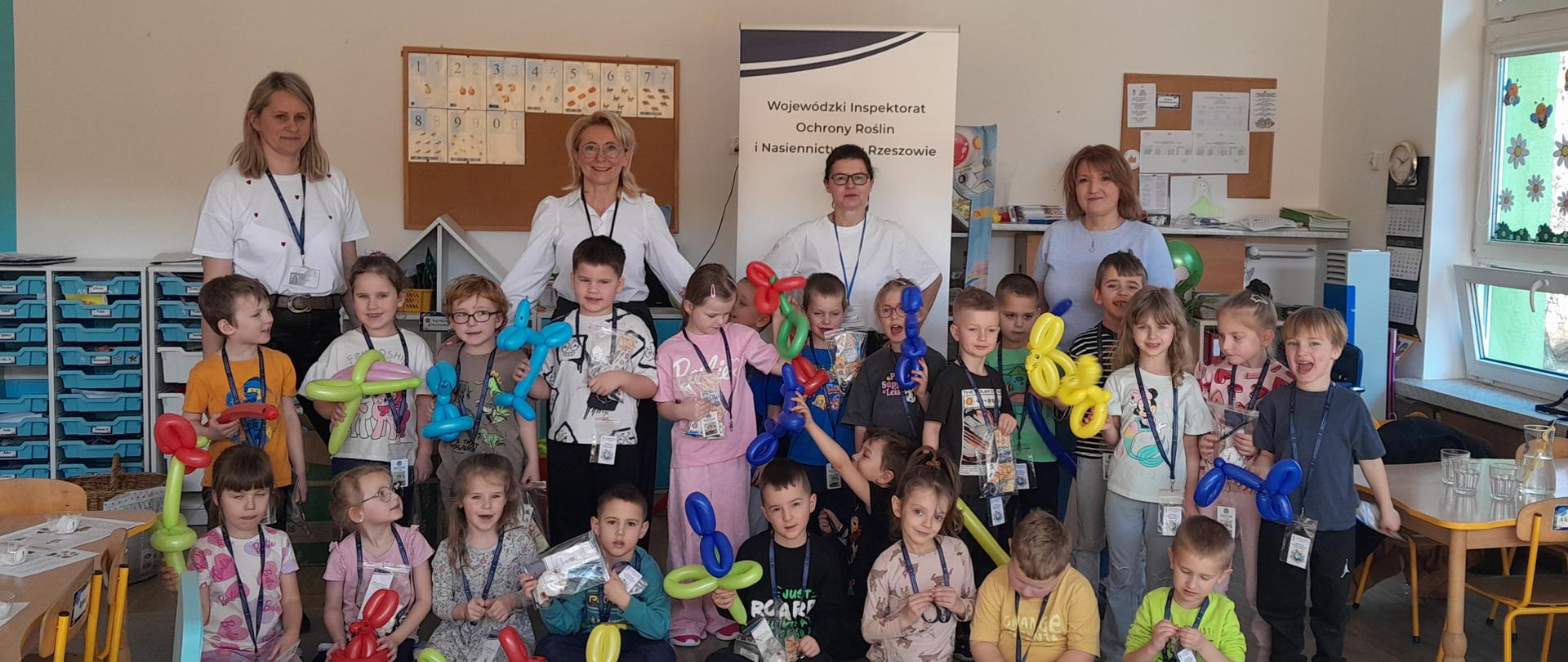 Grupa dzieci przedszkolnych wraz z kilkoma dorosłymi stoi ustawiona do wspólnego zdjęcia w sali. Dzieci trzymają kolorowe balony skręcone w kształty zwierząt oraz drobne upominki w przezroczystych opakowaniach. Dorośli stoją za nimi, uśmiechając się do aparatu. W tle widoczny jest baner z napisem „Wojewódzki Inspektorat Ochrony Roślin i Nasiennictwa w Rzeszowie”, tablice edukacyjne oraz okno z dekoracjami. Atmosfera jest radosna i uroczysta.