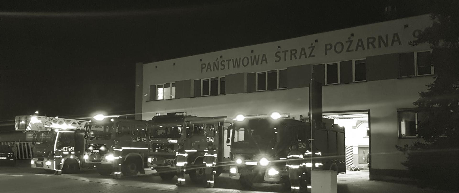 Na zdjęciu widać strażaków przy wozach bojowych, które mają włączone sygnały świetlne