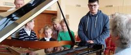 Mężczyzna siedzi przy fortepianie, wokół niego stoją dzieci i młodzież.