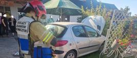 Na zdjęciu stoi strażak PSP obok uszkodzonego samochodu osobowego z otwartą przednią klapą obok osoby postronne