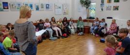 Ok. 20 dzieci siedzi na krzesłach ustawionych w kręgu w dużej sali i słucha prelegentki.