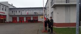 Widok z boku. Plac przy JRG Andrychów. Poczet flagowy wciąga flagę na maszt. Przed budynkiem pododdział złożony ze strażaków zmiany służbowej. Dowódca zalutuje. W tle bramy garażowe. 