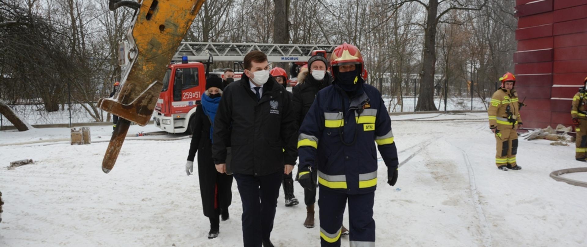 Wojewoda małopolski podąża obok Komendanta Wojewódzkiego PSP. Dookoła śnieg, w oddali strażacy i wóz strażacki.