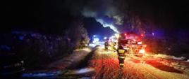 Na zdjęciu w porze nocnej widoczne samochody pożarnicze i strażacy przemieszczający się do pożaru budynku mieszkalnego. Warunki zimowe, śnieg, strażacy w trakcie rozwijania linii gaśniczych.