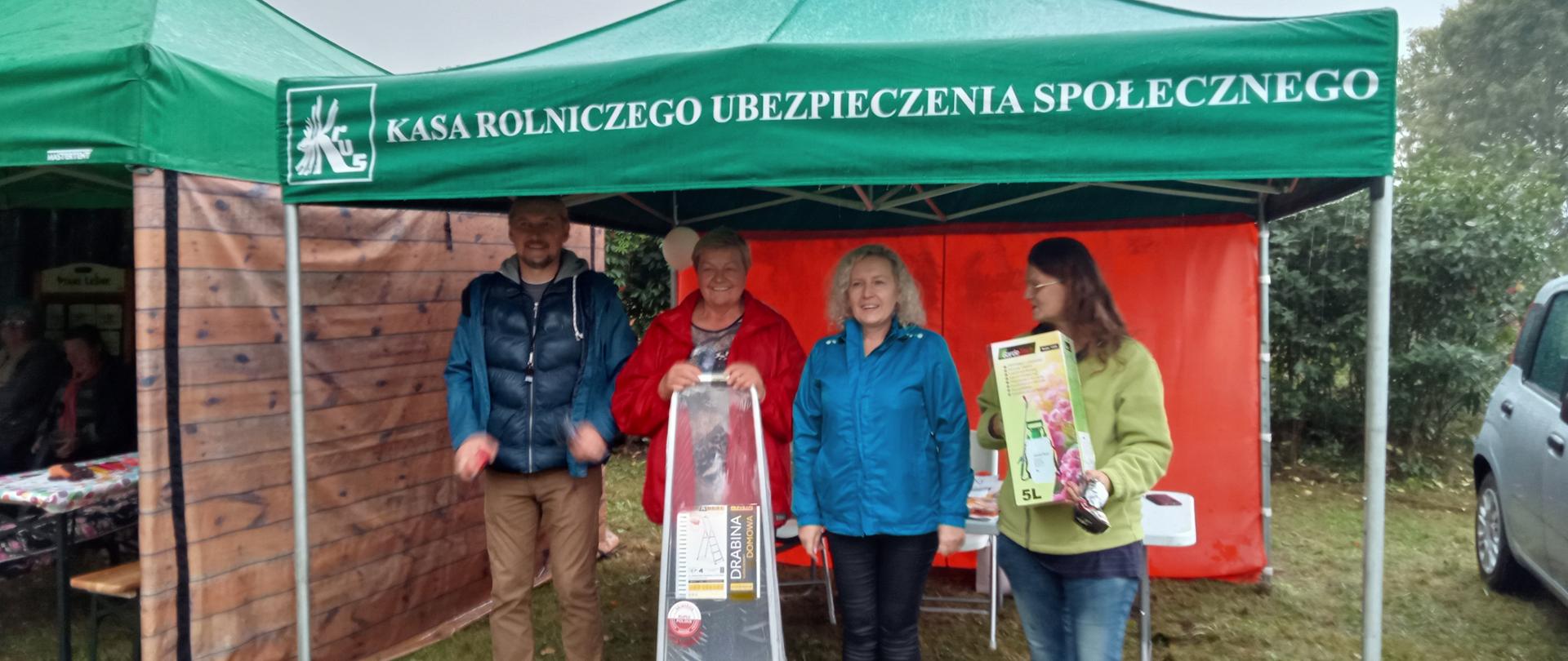 Na środkowym planie zielony namiot stoiska informacyjnego Placówki Terenowej KRUS w Hajnówce. Wewnątrz namiotu stoją cztery osoby, trzy kobiety i jeden mężczyzna. Dwie kobiety trzymają nagrody otrzymane w konkursie.