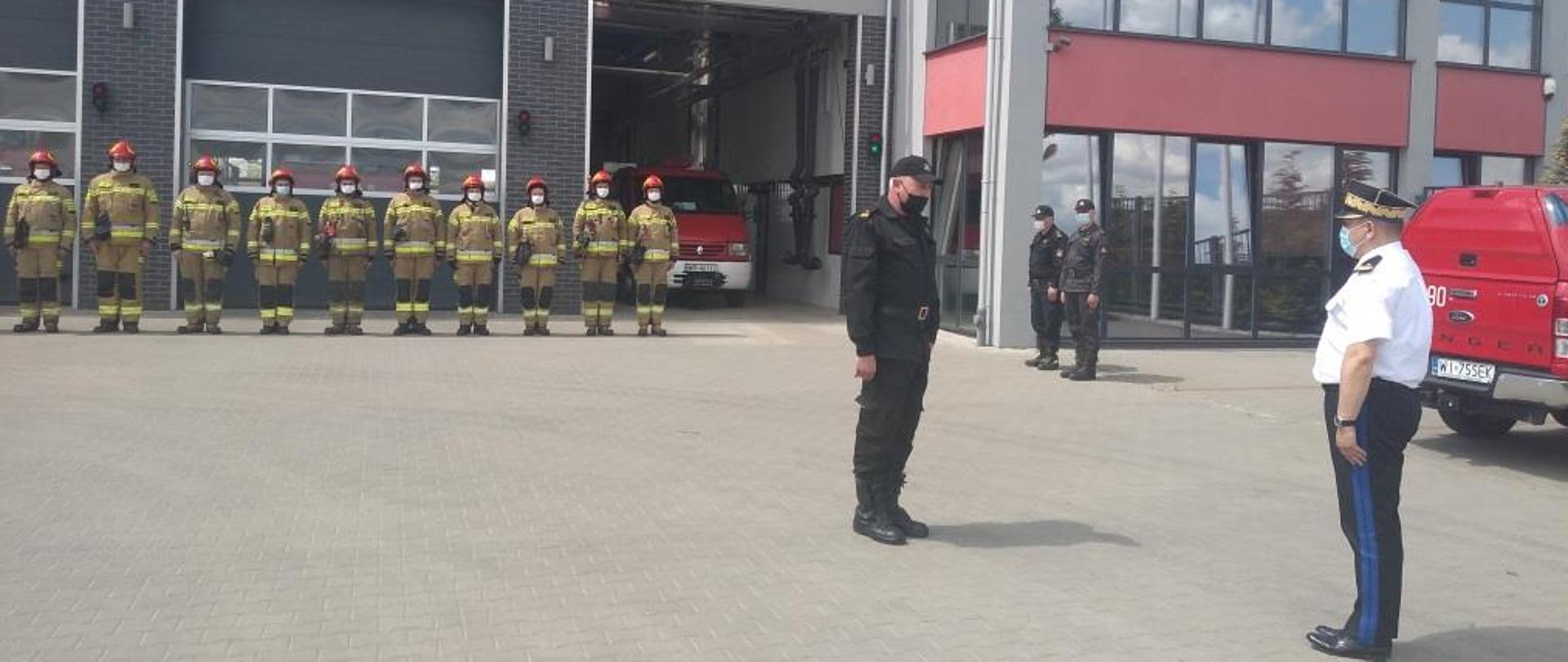 Zdjęcie wykonane przed budynkiem jednostki ratowniczo – gaśniczej Państwowej Straży Pożarnej numer 2 w Radomiu. Przed budynkiem ustawiona w szeregu jest cała zmiana służbowa jednostki. Strażacy ubrani są w ubrania specjalne w kolorze żółtym. Na głowach mają czerwone hełmy. Przed nimi na placu dowódca jednostki składa meldunek Mazowieckiemu Komendantowi Wojewódzkiemu nadbrygadierowi Jarosławowi Nowosielskiemu.