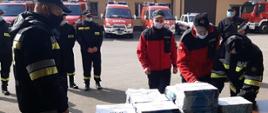 Na zdjęciu, na pierwszym planie strażacy ochotnicy potwierdzają podpisem odbiór ulotek informacyjnych. Przed nimi, na stole leżą pakiety ulotek. Za strażakami widać samochody pożarnicze.