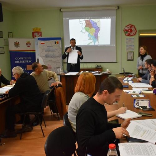 Kilkanaście osób siedzi przy dwóch stołach w trakcie szkolenia, prowadzący pokazuje coś na kartce papieru, W tle dwa rollupy i ekran do prezentacji multimedialnych.