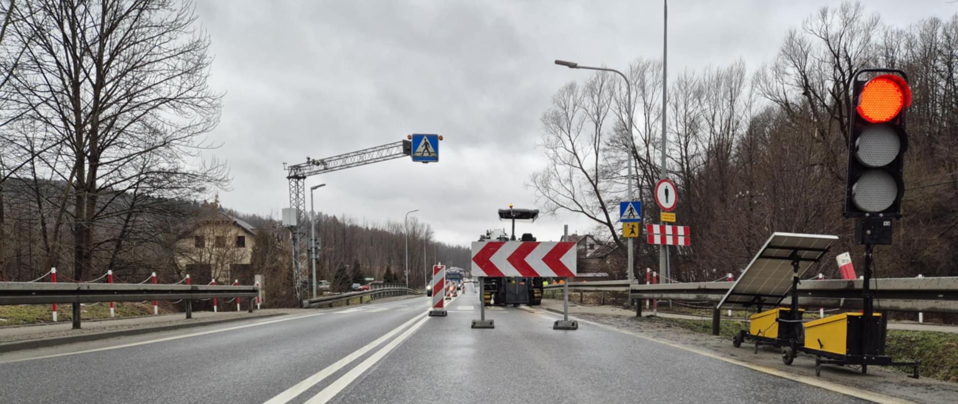 Na zdjęciu widać fragment drogi, na której prowadzone są prace i obowiązuje tymczasowa organizacja ruchu. Jedna z jezdni jest zablokowana dużymi biało‑czerwonymi barierami kierunkowymi. Po prawej stronie stoi przenośna sygnalizacja świetlna zasilana panelem słonecznym – światło jest czerwone. Nawierzchnia jest mokra, a niebo zachmurzone. Po obu stronach drogi rosną drzewa bez liści. W tle widać znaki drogowe, m.in. ostrzegający o przejściu dla pieszych oraz znak zakazu.