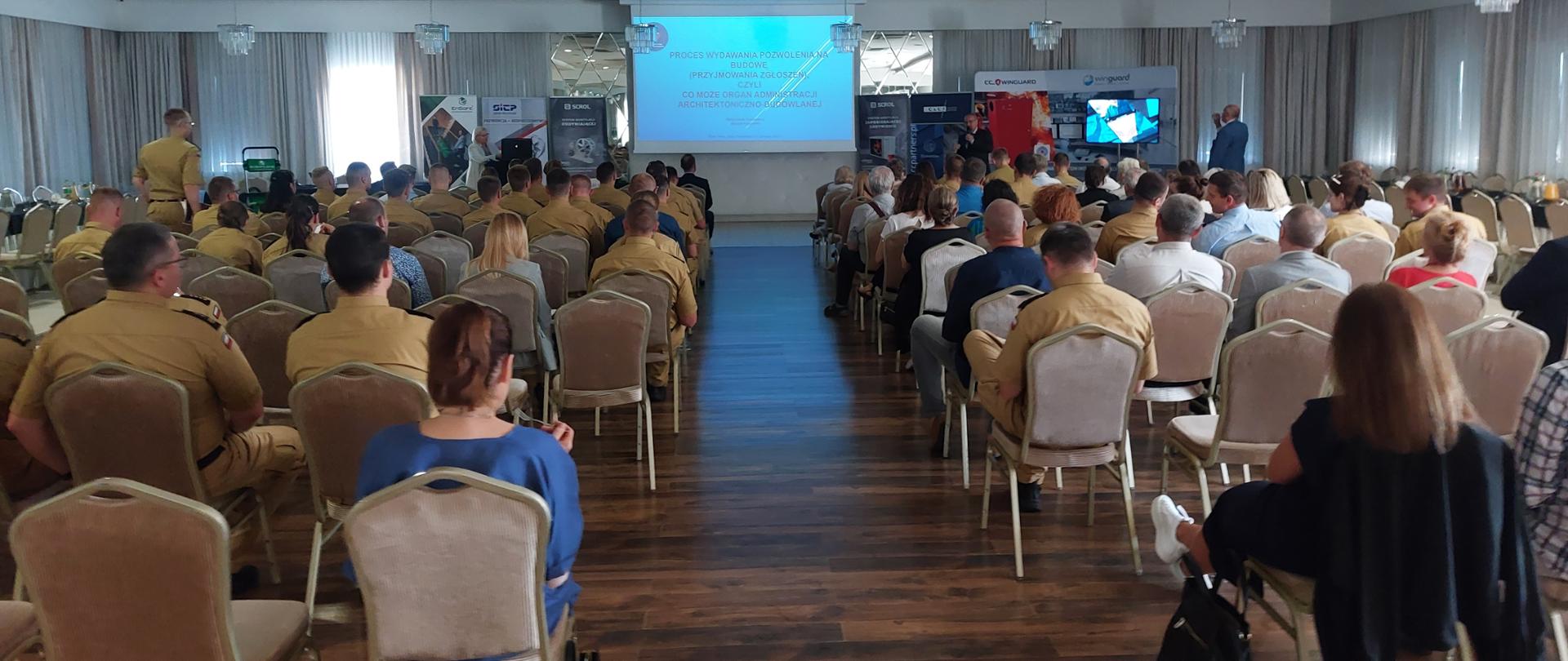 Zdjęcie przedstawia strażaków oraz osoby cywilne podczas seminarium. Osoby siedzą na krzesełkach na sali , natomiast prelegent omawia temat. 