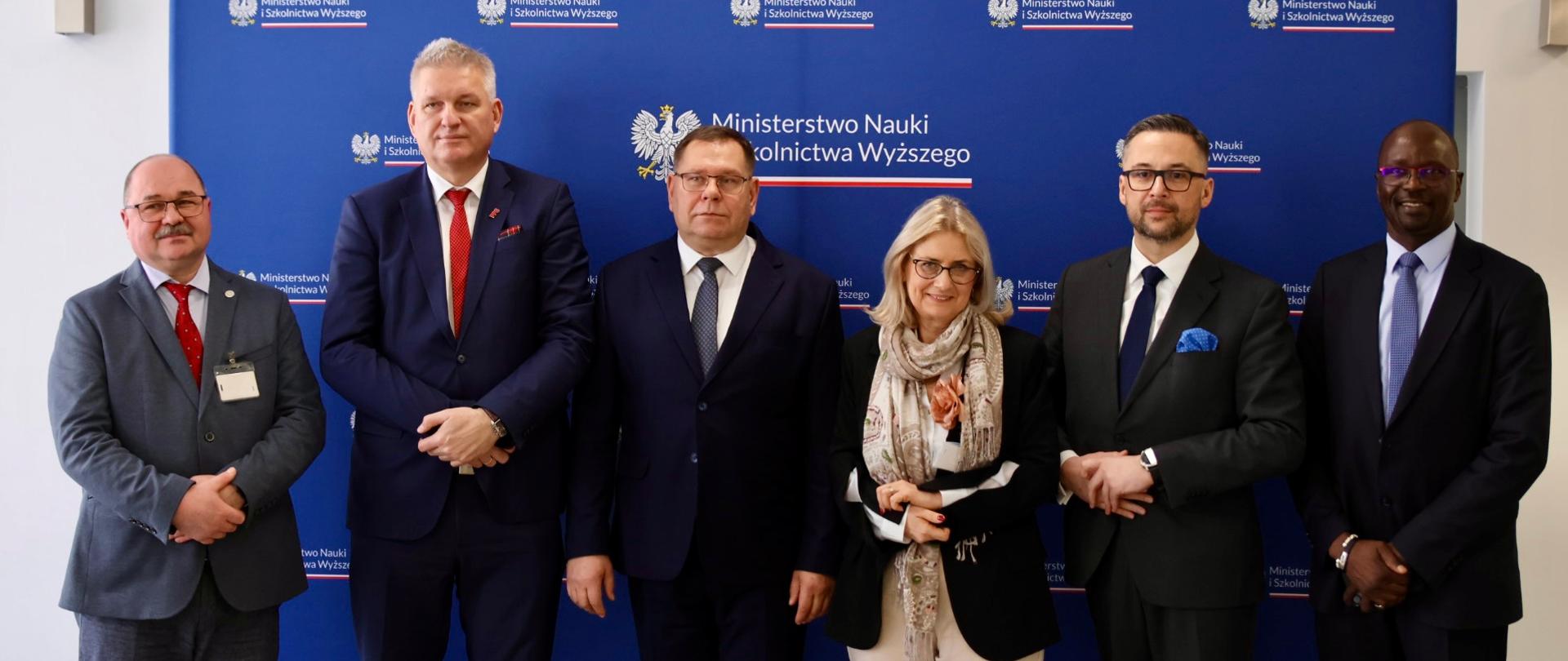 Na tle granatowej ścianki z napisem Ministerstwo Nauki i Szkolnictwa Wyższego stoi sześć osób, drugi z prawej minister Kulasek.