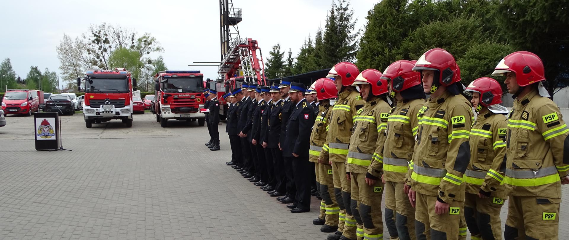 Zdjęcie wykonano w ciągu dnia na placu Komendy Powiatowej Państwowej Straży Pożarnej w Sępólnie Krajeńskim. Przedstawia Strażaków podczas uroczystości obchodów dnia strażaka. Strażacy Państwowej Straży Pożarnej ubrani w mundury wyjściowe i ubrania specjalne stoją w dwuszeregu. W tle mównica i samochody ratowniczo gaśnicze oraz podnośnik hydrauliczny.