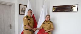 Strażak w mundurze musztardowym ściska dłoń strażaczce w mundurze musztardowym która trzyma czerwoną teczkę za nimi stoją dwie flagi Polski na ścianach wiszą obraz oraz szabla.