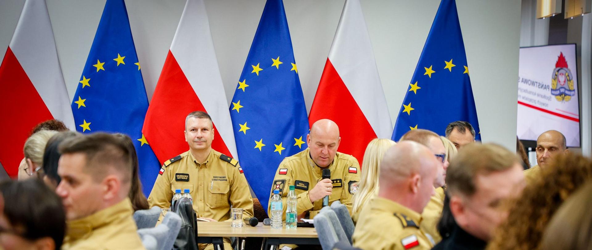 Sala konferencyjna z flagami Polski i Unii Europejskiej w tle. Przy stołach siedzą funkcjonariusze Państwowej Straży Pożarnej w żółtych mundurach służbowych. Jeden z nich mówi do mikrofonu, pozostali słuchają.
