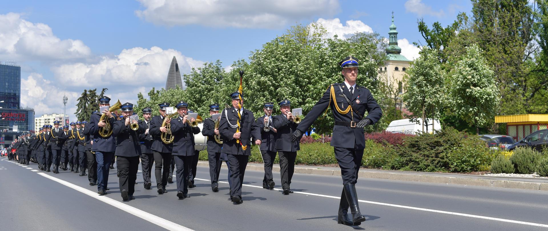 Na zdjęciu widnieje pododdział strażacki w umundurowaniu wyjściowym. Dowódca pododdziału na jego czele, następnie za nim orkiestra dęta OSP oraz kolejne pododdziały. Wszyscy maszerują asfaltową aleją płk. Cieplińskiego w Rzeszowie, w tle zieleń miejska i zabudowania.