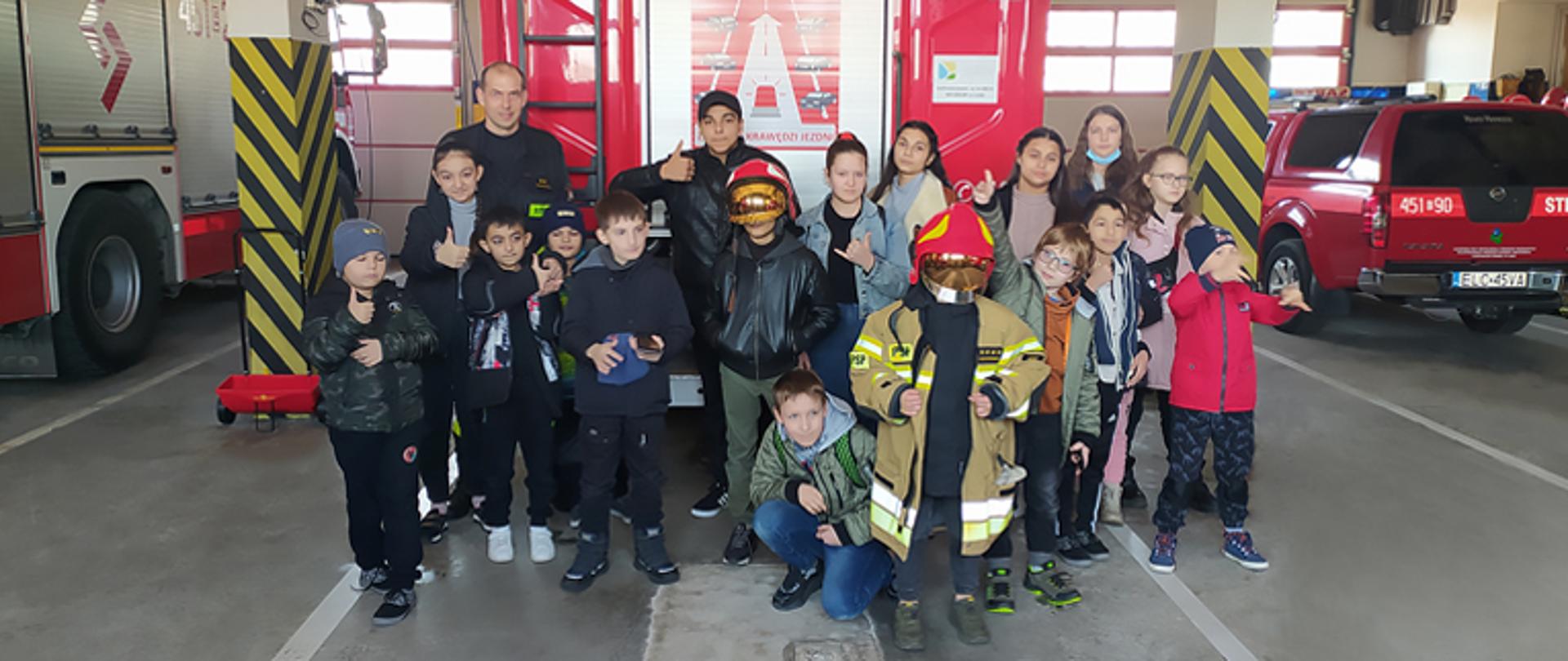 Na pierwszym planie w garażu grupka dzieci z Ukrainy pozują do fotografii, unoszą do góry prawy kciuk, jeden chłopiec na środku kadru ubrany jest w kurtkę bojową strażaka koloru jasnożółtego oraz ma założony hełm barwy czerwonej na głowie, w tle za nimi stoi auto ciężarowe barwy czerwonej, po lewej i prawej stronie samochody bojowe straży pożarnej