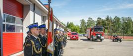 
Na zdjęciu pododdziały strażaków w mundurach wyjściowych koloru czarnego oraz w ubraniach specjalnych koloru musztardowego z hełamami koloru czerwonego, stojące w dwuszeregu na tle zaparkowanych pojazdów i budynku.