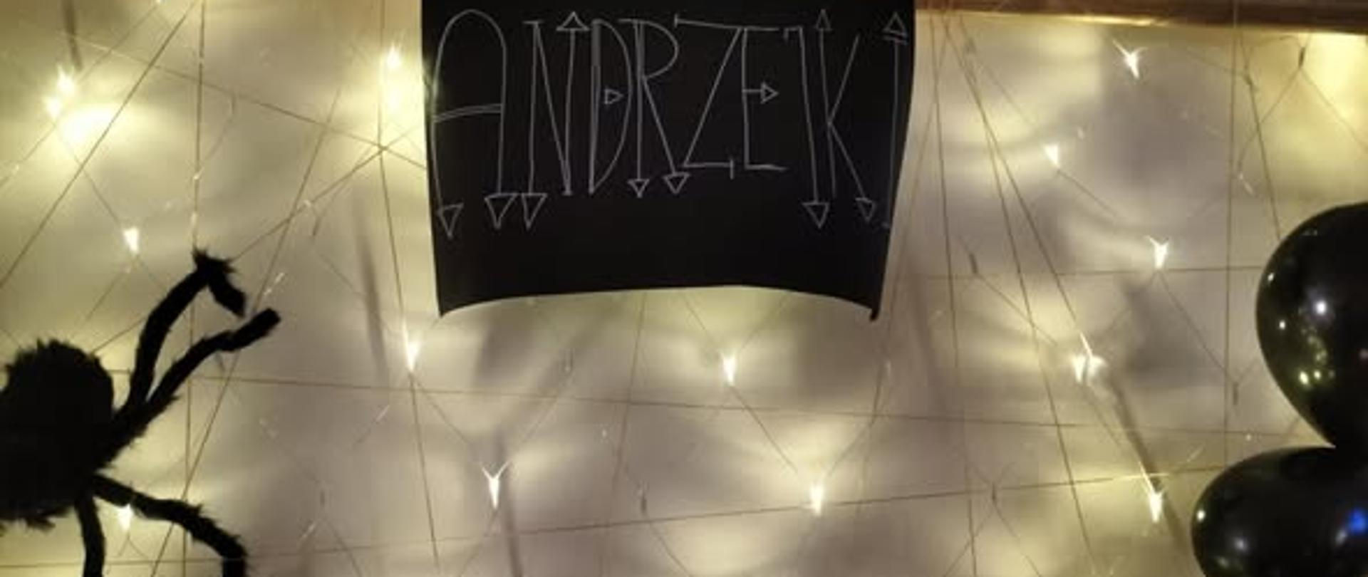 zdjęcie drewnianej ramki zawieszonej na ścianie, wzdłuż i wszerz rozciągnięte światełka. Po środku ramki na górze czarny karton z napisem Andrzejki. Po lewej stronie czarny pająk z materiału.