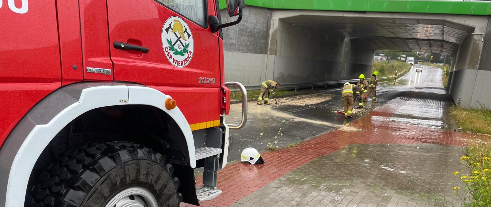Widać czerwony samochód strażacki z oznaczeniami OSP Wierzbica stojący na brukowanym chodniku. W tle, pod wiaduktem, kilku strażaków w żółtych hełmach i ubraniach bojowych usuwa skutki zdarzenia – na jezdni leżą trociny i woda. Na pierwszym planie leży biały hełm strażacki.