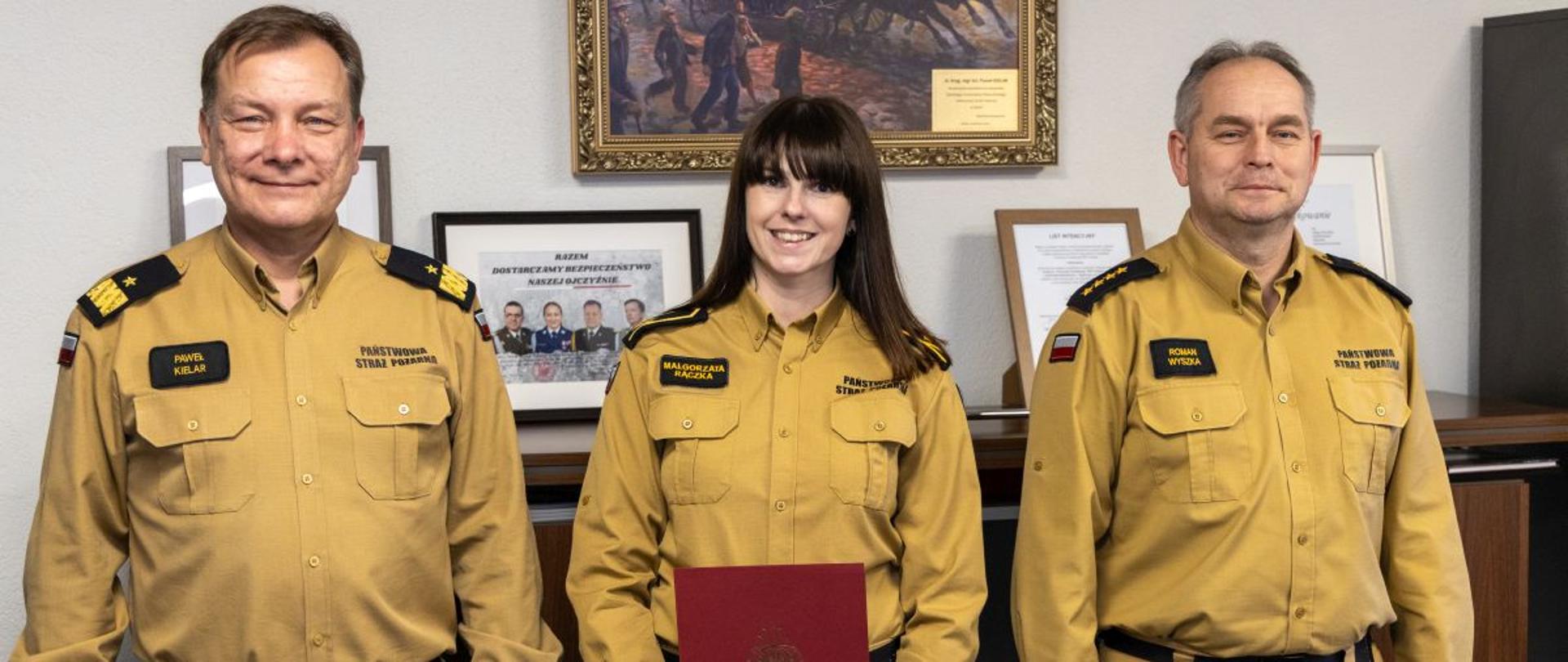 Mł. asp. Małgorzata Rączka prezentuje czerwony folder z aktem mianowania na stanowisko psychologa w Wydziale Planowania Operacyjnego. Obok niej stoją nadbryg. Paweł Kielar i st. kpt. Roman Wyszka. Tło stanowi oficjalna przestrzeń biurowa z certyfikatami i obrazem batalistycznym.