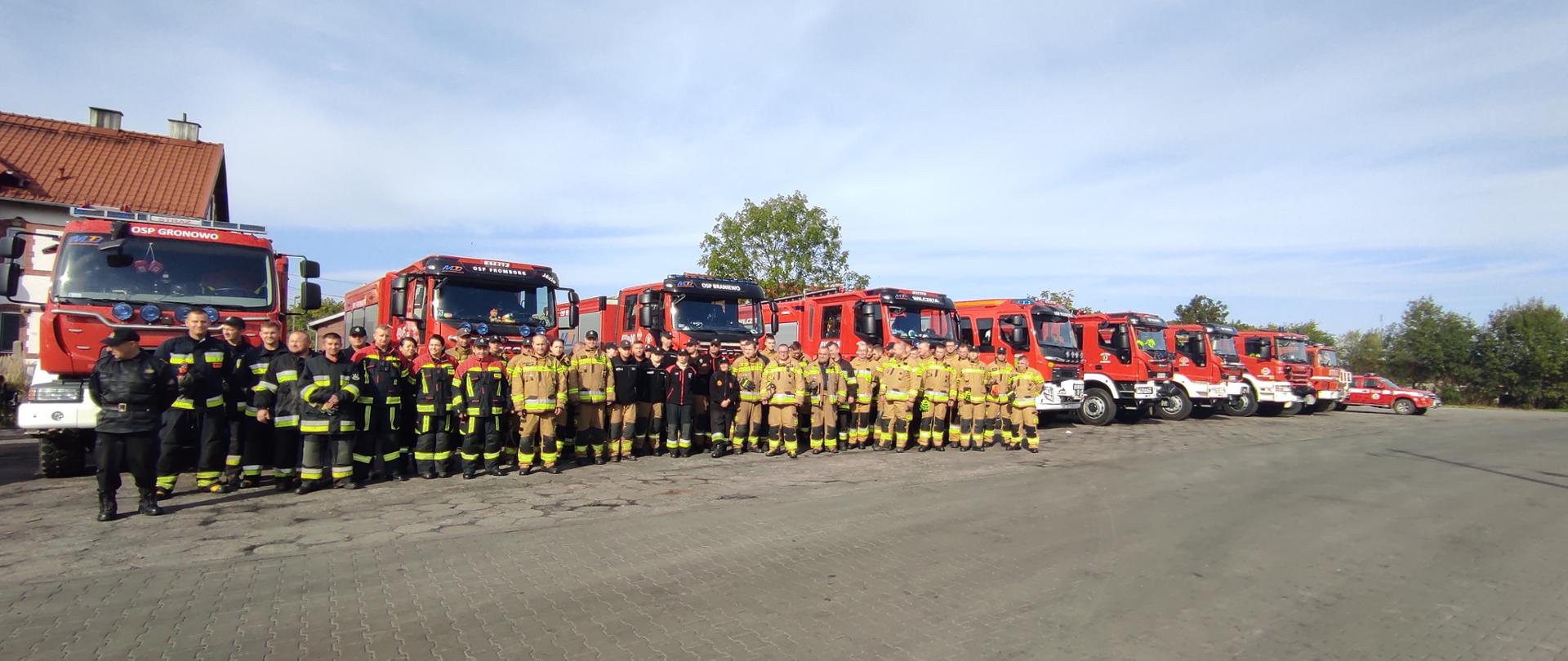 Na dużym placu ustawiło się kilkudziesięciu strażaków w mundurach. Za nimi kilkanaście dużych samochodów strażackich. Pochmurne niebo.