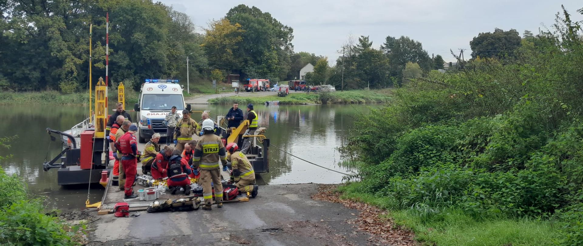 Na promie na rzece Odrze stoi karetka pogotowia. Przed nią ratownicy z zespołu ratownictwa medycznego i Lotniczego Pogotowia Ratunkowego wspólnie ze strażakom udzielaj pomocy poszkodowanej osobie. W tle rzeka Odra , a po drugiej stronie rzeki ustawione są pojazdy pożarnicze. 