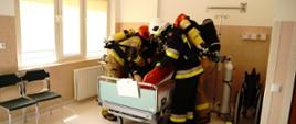 Strażacy w ubraniach specjalnych w hełmach strażackich ewakuujący pacjenta szpitala z sali. Przygotowują pacjenta do transportu na noszach wraz z kardiomonitorem przenośnym, butlą z tlenem i pompą infuzyjną. 
