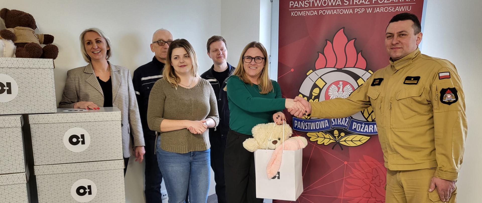 Na zdjęciu widzimy moment przekazania pluszowych misiów w sali konferencyjnej w budynku Komendy Powiatowej PSP w Jarosławiu. Po lewej znajdują się pudełka z logo O-I (Owens-Illinois), a w centralnej części przedstawiciele Huty Szkła Jarosław, w tym Pani Monika Falandysz, która przekazuje w torbie prezentowej Misia Ratownika Zastępcy Komendanta Powiatowego PSP w Jarosławiu. W tle ustawiono roll-up KP PSP w Jarosławiu.