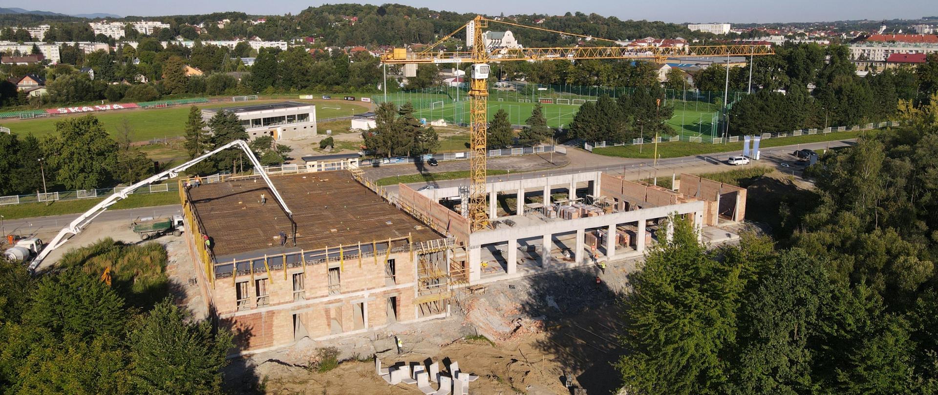 Widok z góry. Plac budowy nowej komendy straży pożarnej. Na środku dźwig, z lewej strony trwa wylewanie stropu. W tle zabudowania miejskie i wzgórza. 