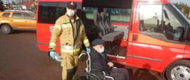 Zdjęcie przedstawia strażaka i osobę starszą na wózku inwalidzkim, która strażacy przetransportowali do punktu szczepień.