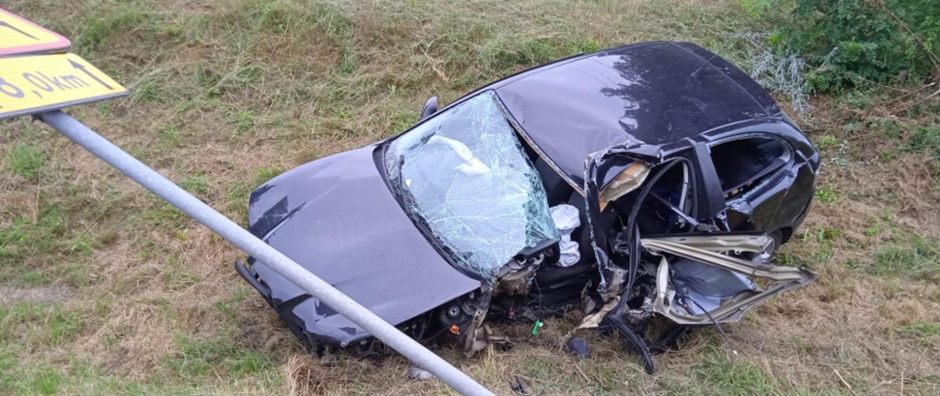 Na zdjęciu widoczny rozbity samochód osobowy marki Seat koloru ciemnego. Zdjęcie wykonane od strony lewego przodu pojazdu. Widoczny także pochylony znak drogowy koloru żółtego.