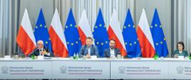 Sala konferencyjna Ministerstwa Spraw Wewnętrznych i Administracji, na tle ustawionych naprzemiennie flag Polski i Unii Europejskiej siedzi kilka osób przy stole prezydialnym. Na stole widnieje nazwa ministerstwa oraz herb Polski, a przed uczestnikami znajdują się mikrofony, dokumenty i butelki wody.