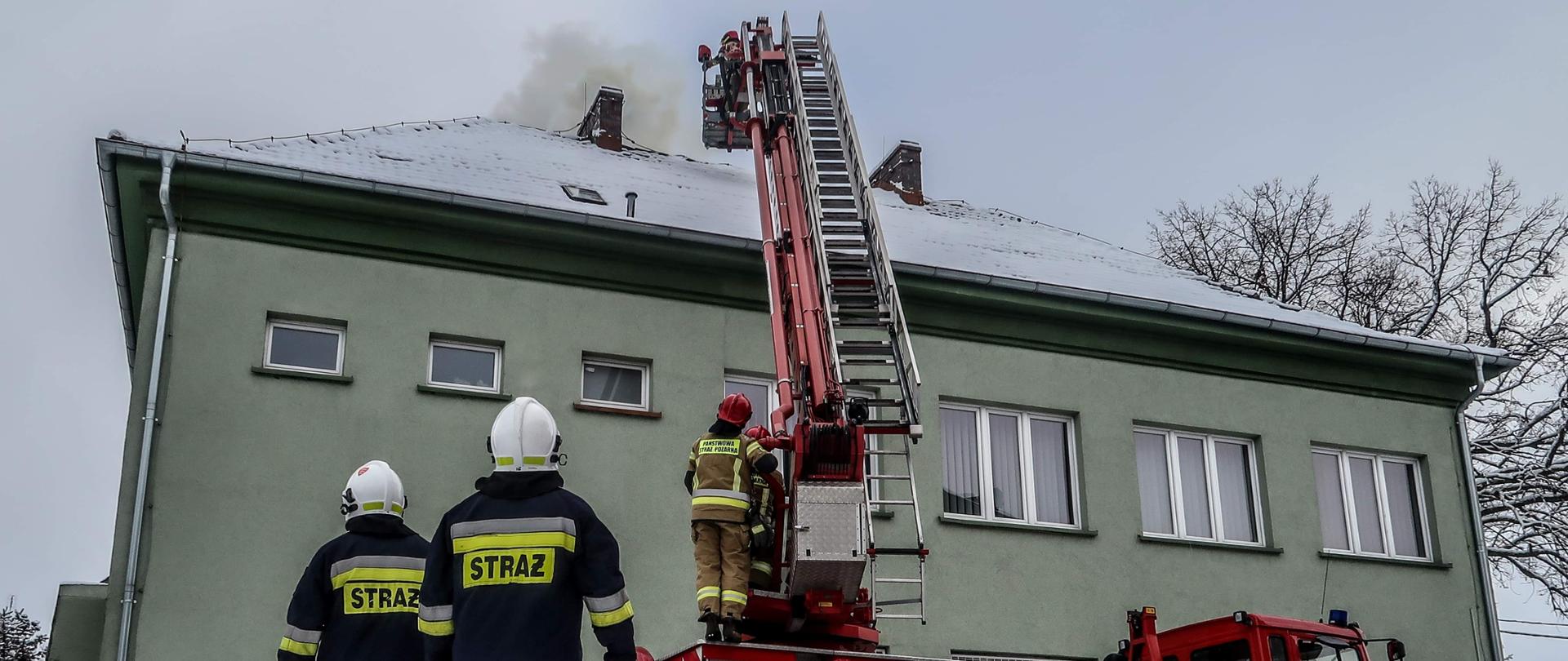 Rozstawiony podnośnik hydrauliczny. W koszu strażacy gaszący komin. Na pierwszy planie dwóch ochotników.