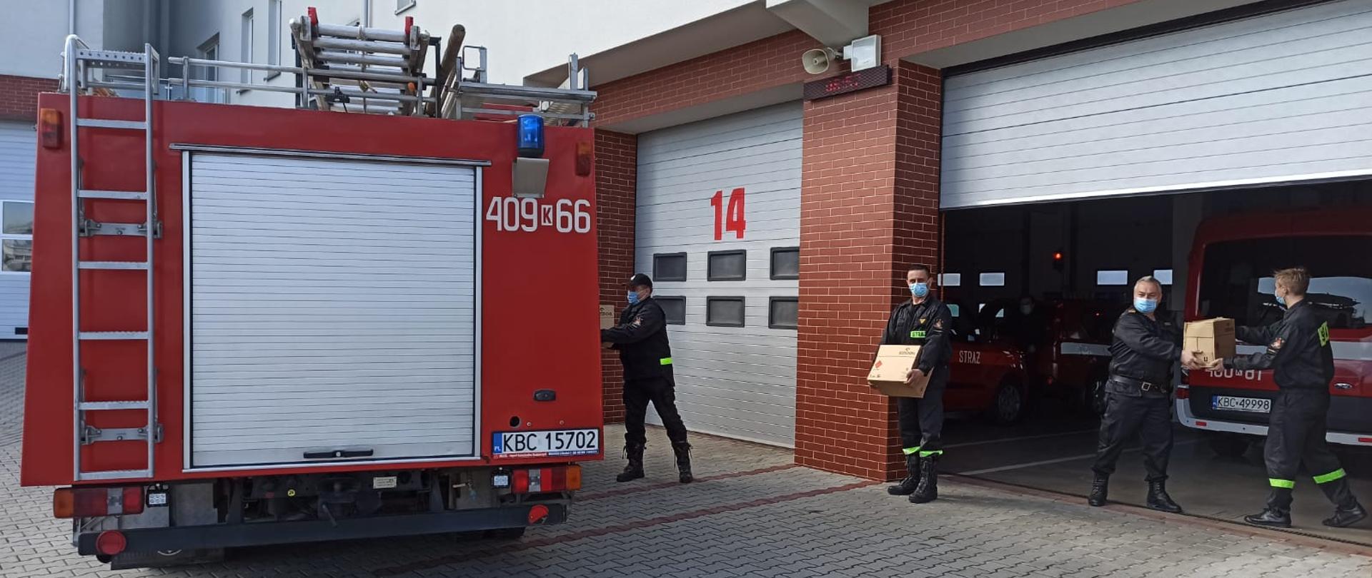 Czterech mężczyzn ubranych w czarny mundur strażacki z odblaskowymi paskami, czarnymi butami, na twarzy ma ubrana maseczkę chirurgiczną koloru biało niebieskiego, stoją na zewnątrz garażu przeznaczonego na pojazdy pożarnicze. Obok znajduje się samochód pożarniczy koloru czerwonego z napisem koloru białego 400-66 oraz na dachu ma drabinę przenośną. Mężczyźni stoją w odległości każdy od siebie ok. 2 metry i podają sobie z rak do rak pojemniki kartonowe. Jeden z mężczyzn wkłada do pojazdu pożarniczego pojemnik kartonowy. W tle jest zamknięta brama garażowa koloru białego. Budynek w kolorze białym natomiast do wysokości pierwszego piętra są płytki koloru ceglanego