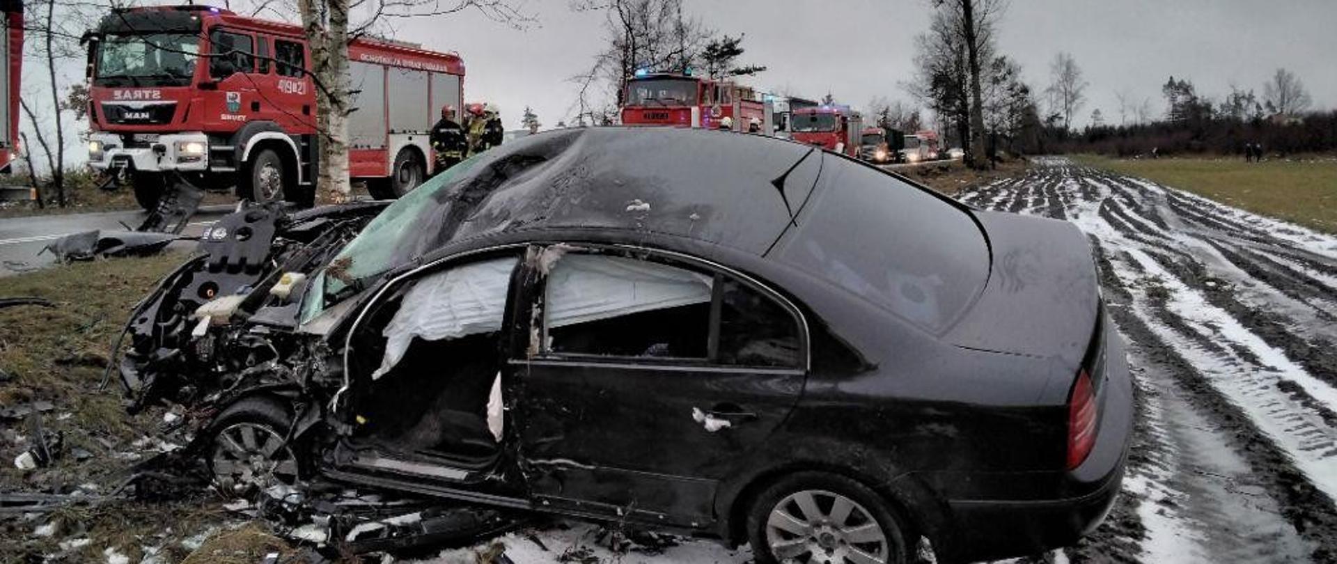 Czarny samochód z mocno rozbitym przodem oraz wystrzelonymi poduszkami powietrznymi stoi na poboczu drogi w polu. W oddali widać samochody strażackie uczestniczące w działaniach ratowniczych.