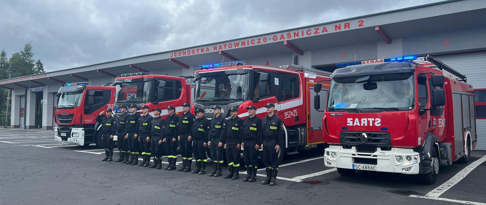 Na zdjęciu widoczny budynek komendy państwowej straży pożarnej, nad garażami napis: jednostka ratowniczo-gaśnicza numer dwa. Przed budynkiem stoją w szeregu strażacy w czarnych ubraniach koszarowych. Za nimi ustawione przed bramami garażowymi cztery czerowne samochody specjalne.